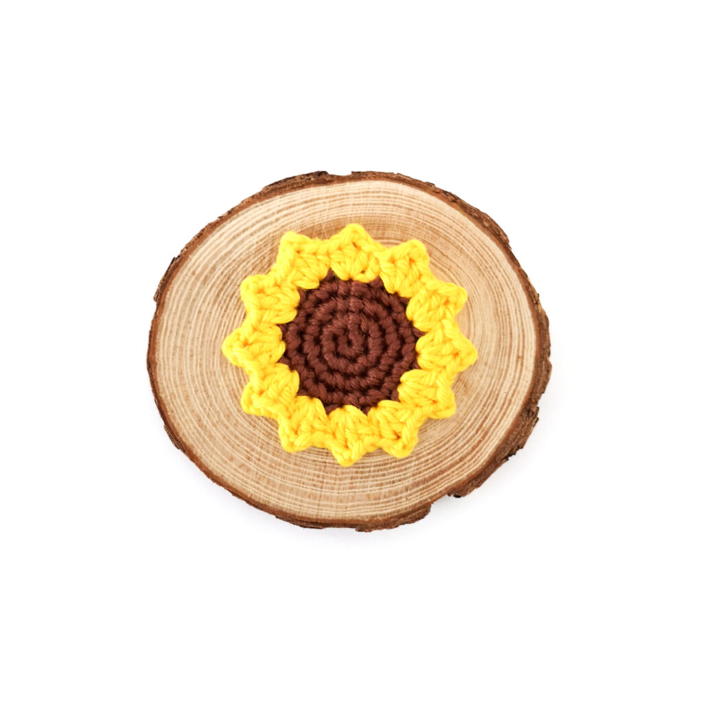 A small yellow crocheted sunflower lying on a round wood slice against a white background.