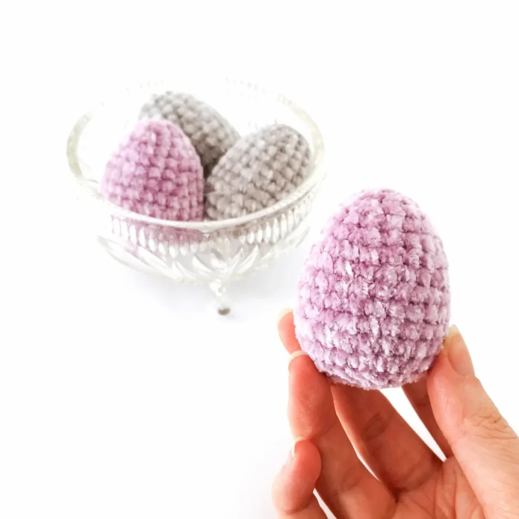 A crochet purple velvet egg being held up in front of a bowl of purple and grey crocheted eggs.