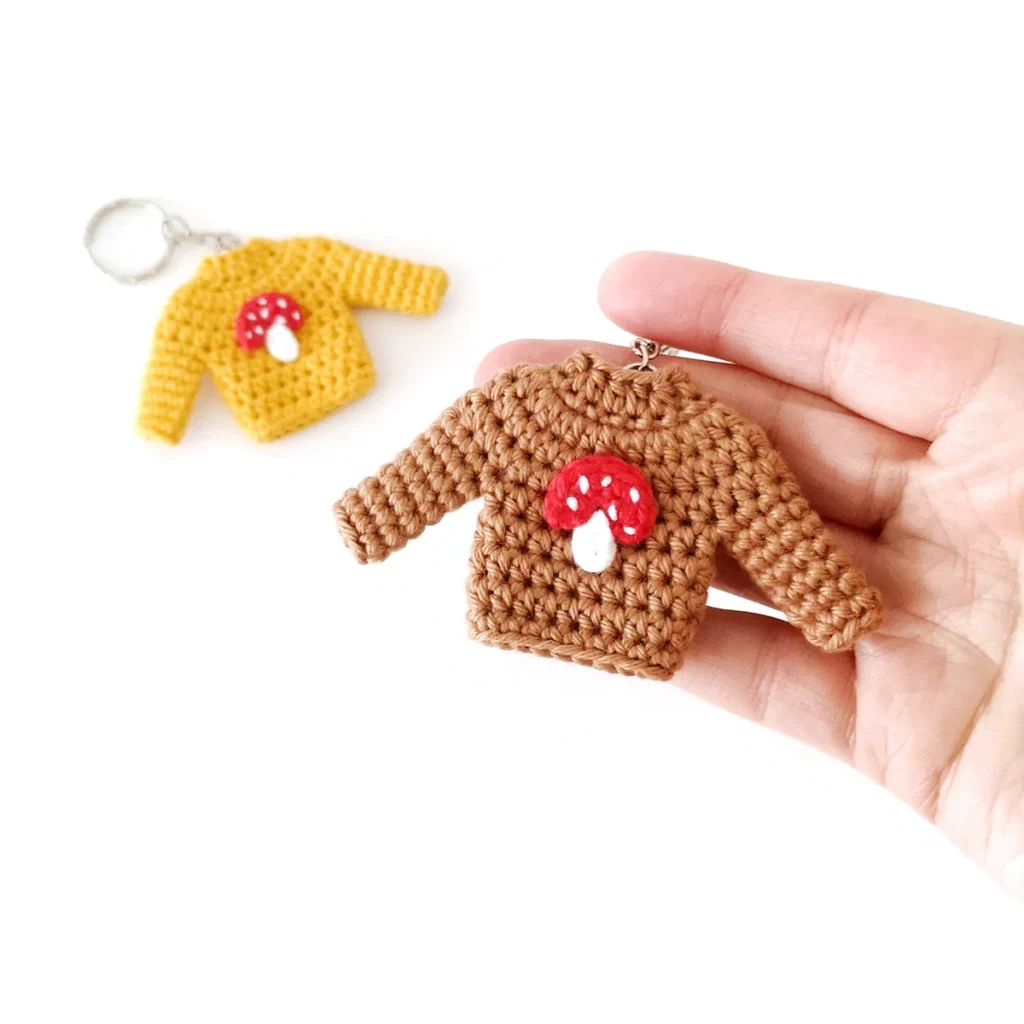 A brown mini crochet sweater keychain with a red and white toadstool applique on the front, being held up by a hand, with a yellow crochet sweater keychain in the background..