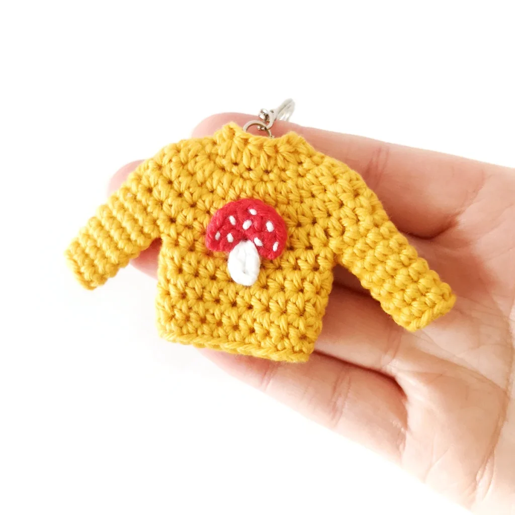 A yellow mini crochet sweater keychain with a red and white applique on the front, being held up by a hand.