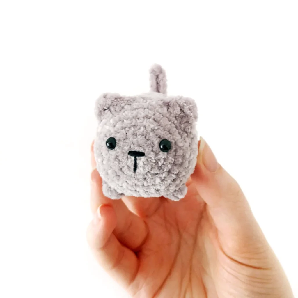 Small plush grey crochet cat being held up by a hand against a white background.
