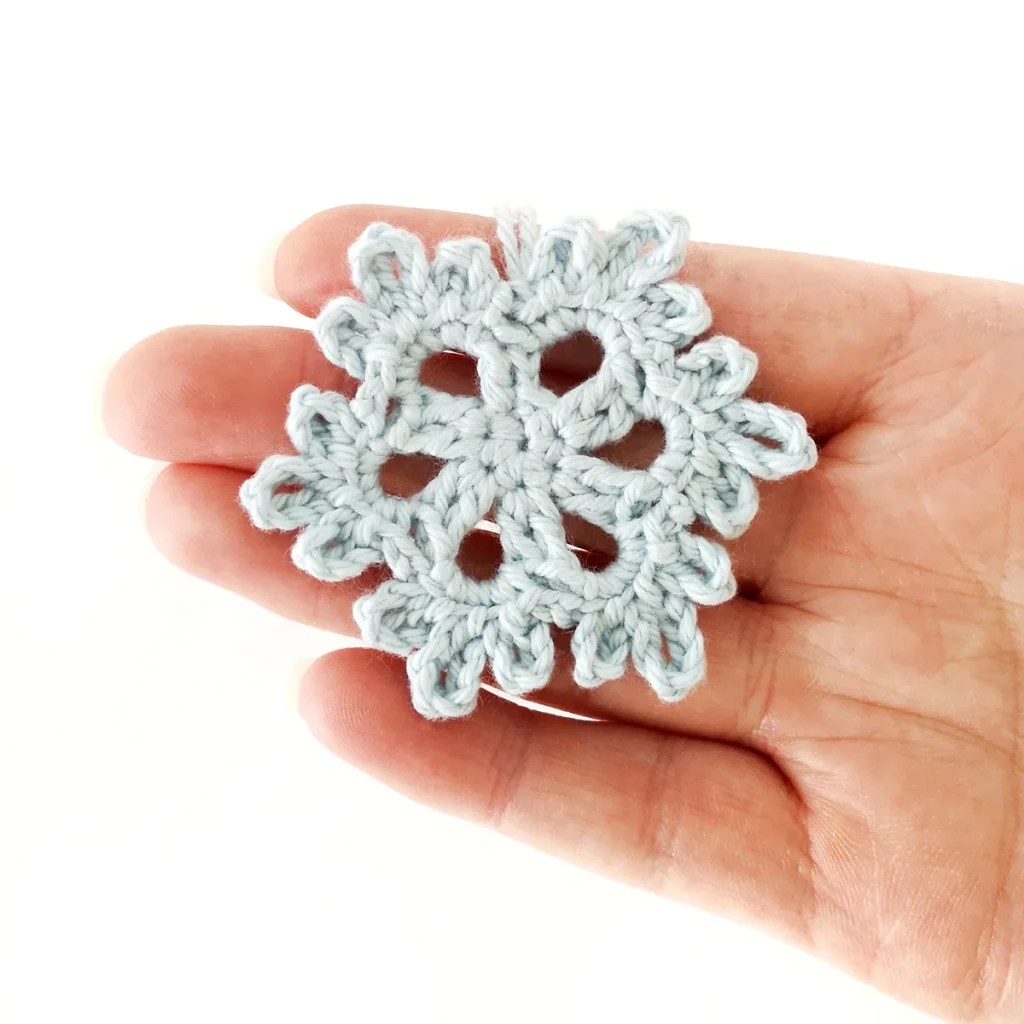 A light blue crochet snowflake being held up on the fingers of a hand.