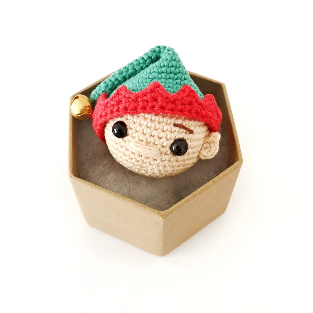 Crochet elf bauble wearing a green Santa hat with red trim and a gold bell sitting in a small hexagonal brown box.
