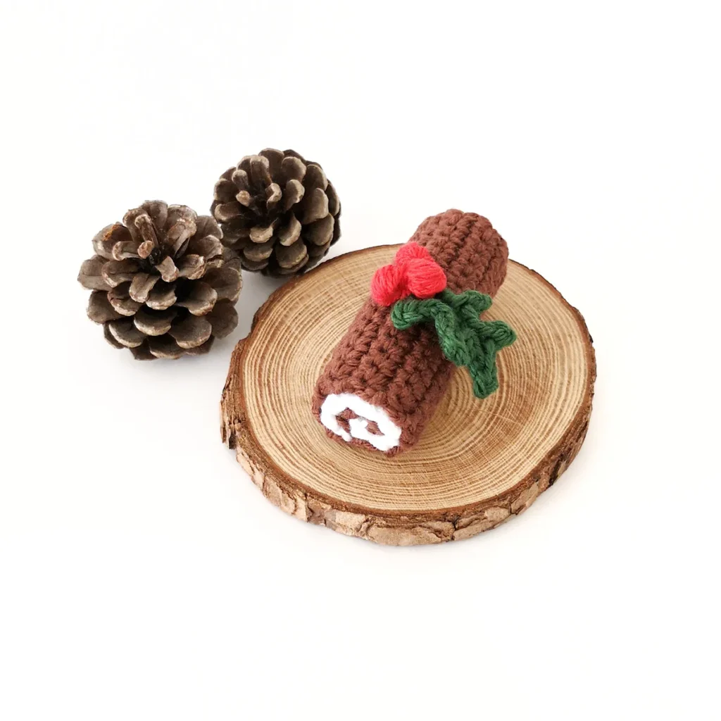 Crochet yule log with holly adornment sitting on a wood round next to two pine cones.
