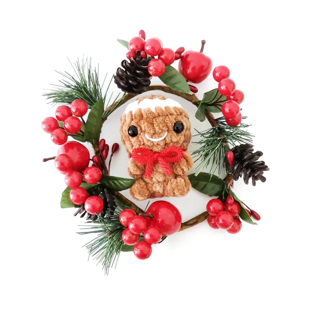 A crochet gingerbread man plush toy lying in the centre of a garland of leaves, pine cones and red berries.