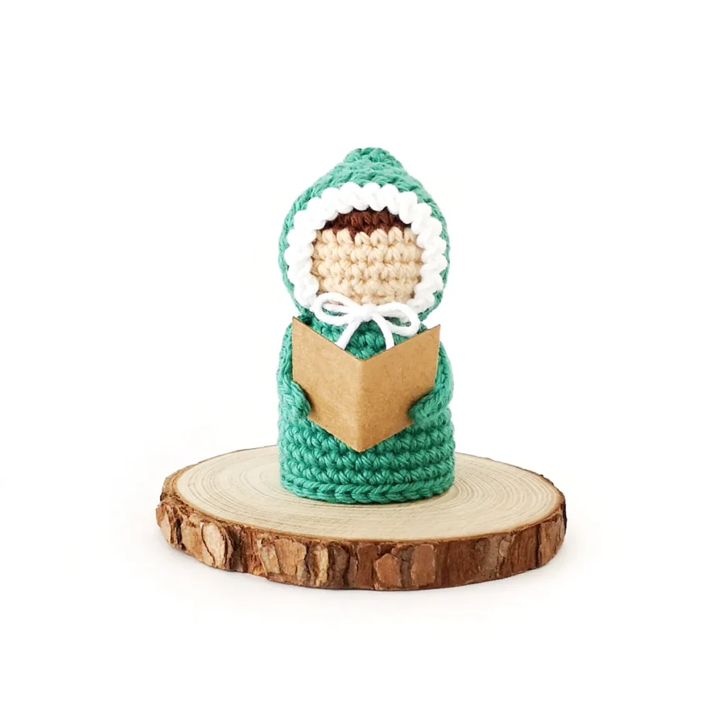 A crochet carol singer, wearing a green outfit with white trim, standing on a wood round, holding an open book.