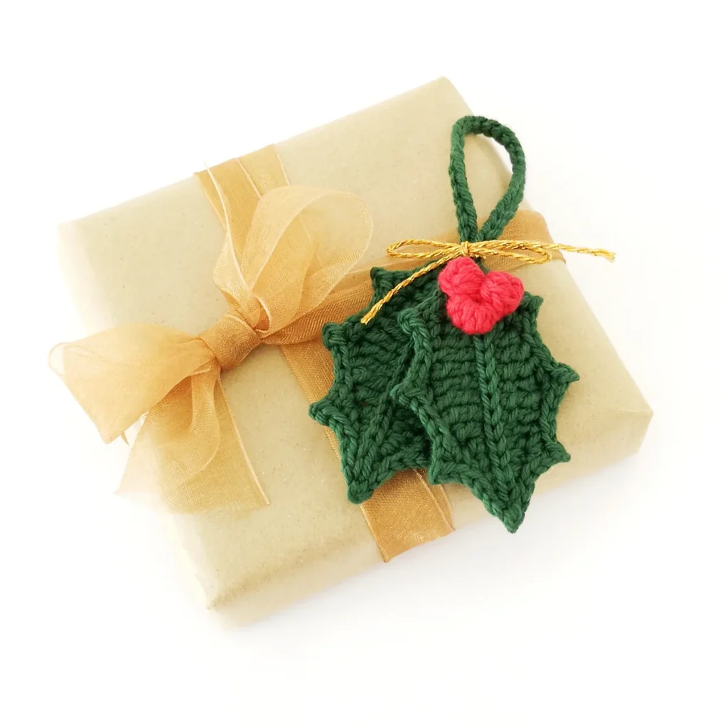 Two crochet holly leaves connected by a hanging loop tied with gold yarn with a cluster of red berries, lying on a brown paper wrapped gift.