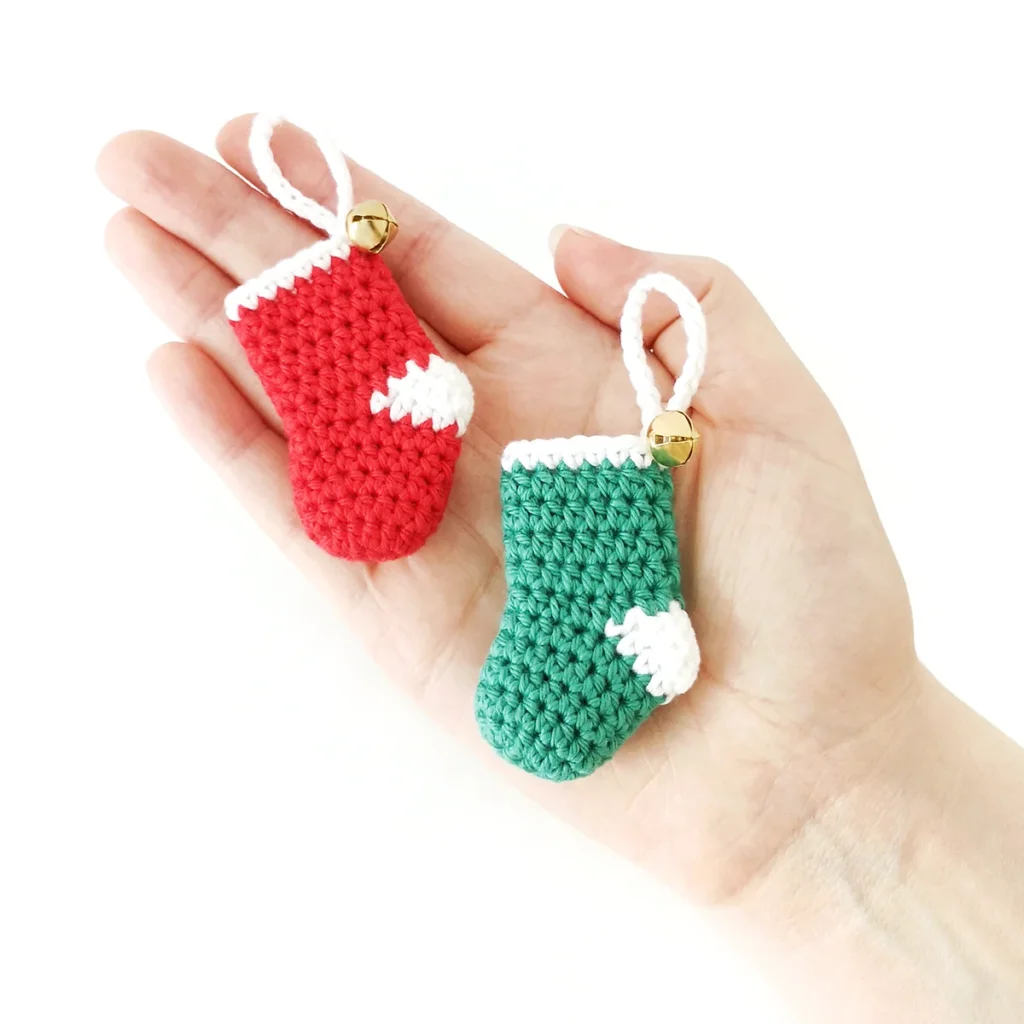 Two small crochet Christmas stockings, one red and one green adorned with a gold bell, resting on the palm of a hand.