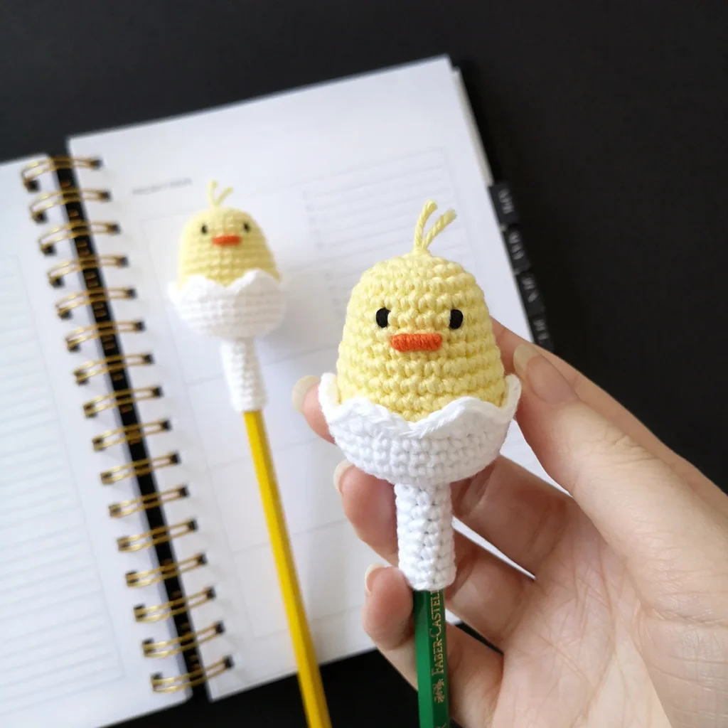 A small yellow crocheted chick in a white half shell atop a green pencil being held up with another chick pencil topper in the background lying on top of an open notebook.