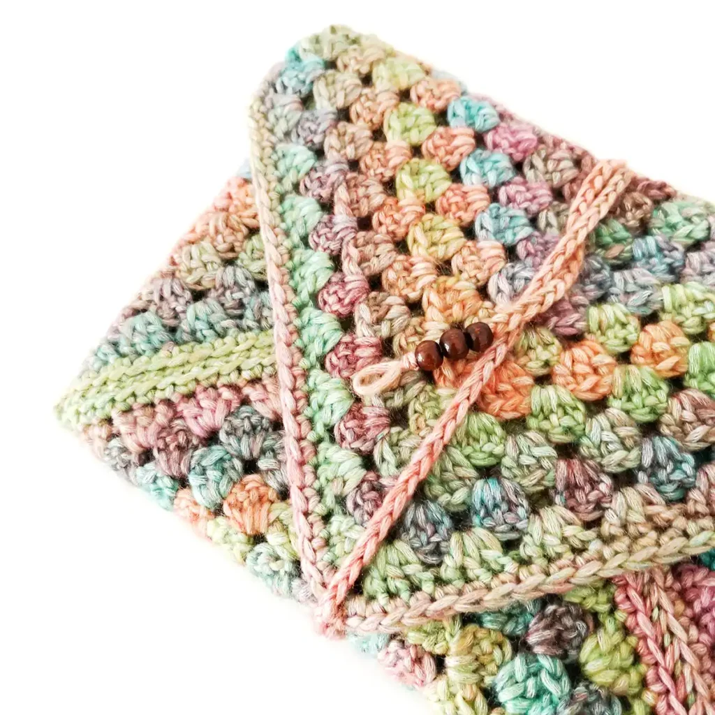 A close up image of a closed multicoloured crochet book sleeve with a beaded tie on a white background.