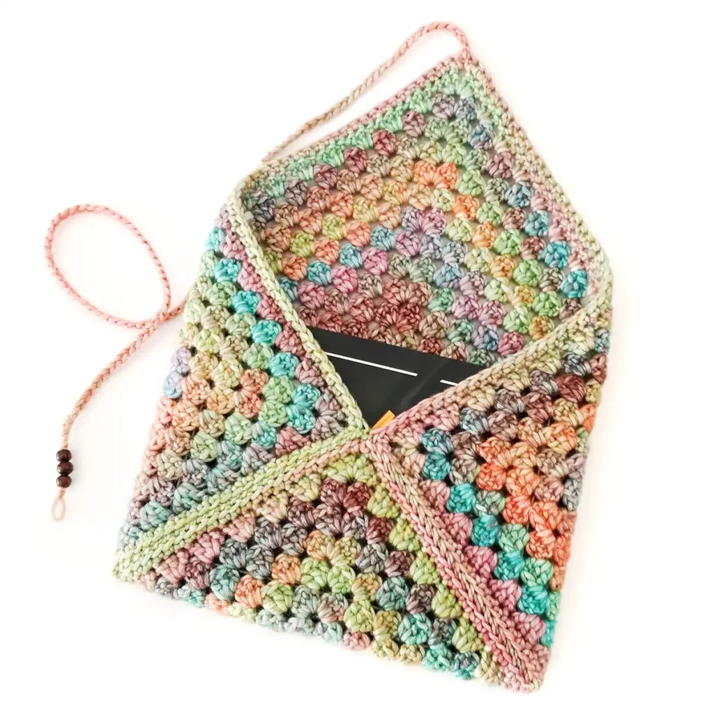 A multicoloured granny square crochet book sleeve with a triangular flap and beaded tie, holding a black book inside, lying on a white background.