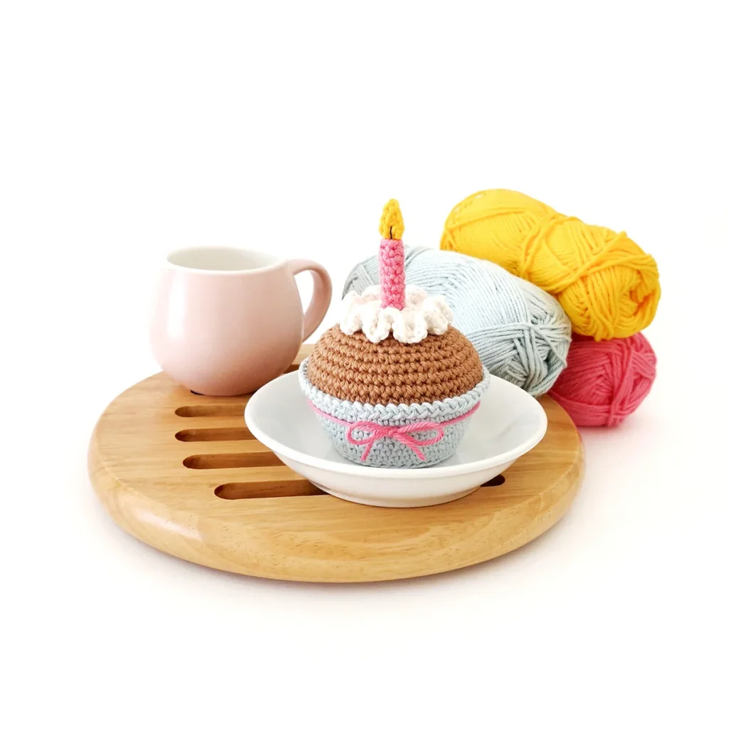 A crochet cupcake with white wavy icing,  a pink candle with a yellow flame and a light blue cupcake liner tied with a pink bow. The cupcake is sitting on a small white dish on a round wood mat next to a small pink teacup with light blue, yellow and pink yarn behind it.