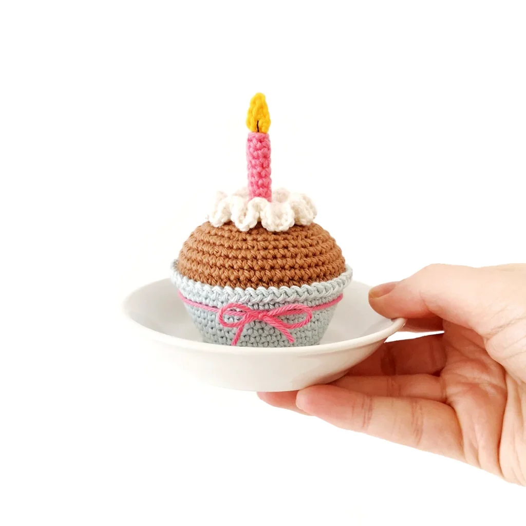 A crochet cupcake with white wavy icing,  a pink candle with a yellow flame and a light blue cupcake liner tied with a pink bow. The cupcake is sitting on a small white dish held in one hand.