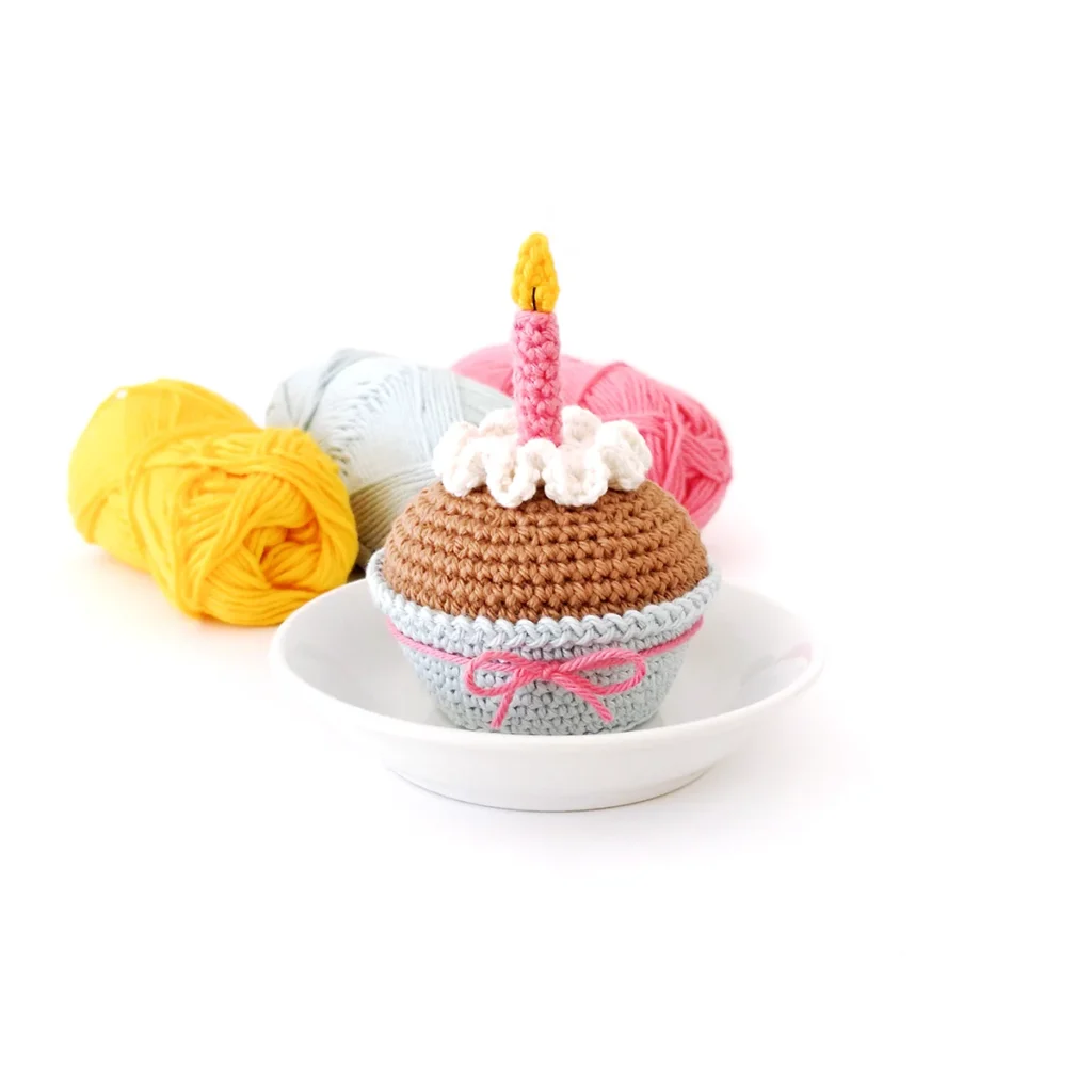 A crochet cupcake with white wavy icing,  a pink candle with a yellow flame and a light blue cupcake liner tied with a pink bow. The cupcake is sitting on a small white dish with 3 skeins of yarn in the background in the colours yellow, light blue and pink.