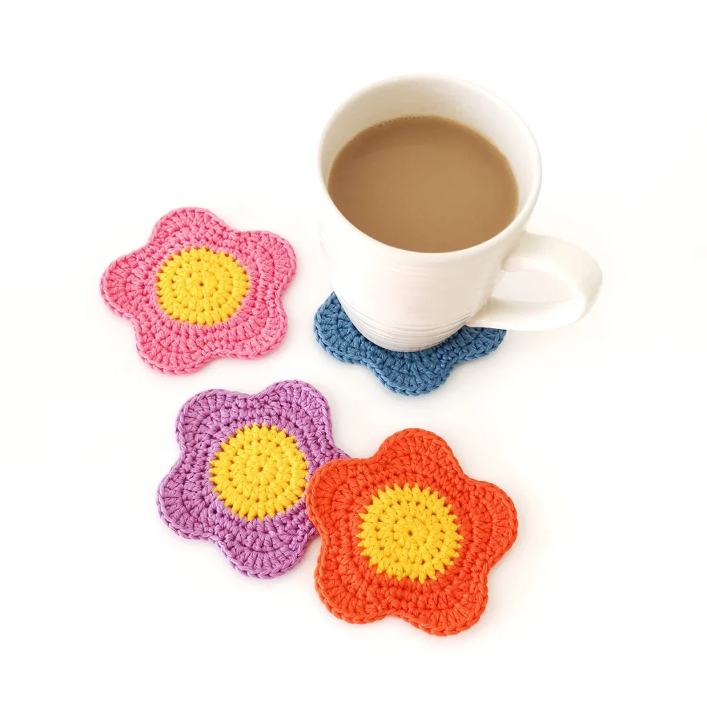 Four crochet flower coasters in pink, purple, orange, and blue with yellow centers. A white mug filled with coffee is sitting on the blue flower coaster.