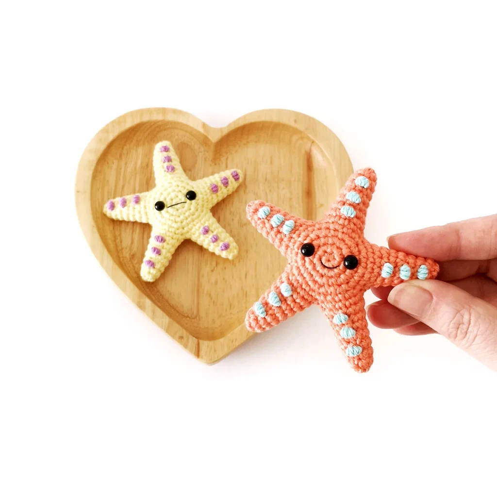 Two small crochet starfish, one orange with blue spots and one yellow with purple spots. The yellow starfish sits on a heart-shaped wooden dish, while a hand holds the orange starfish in the foreground.