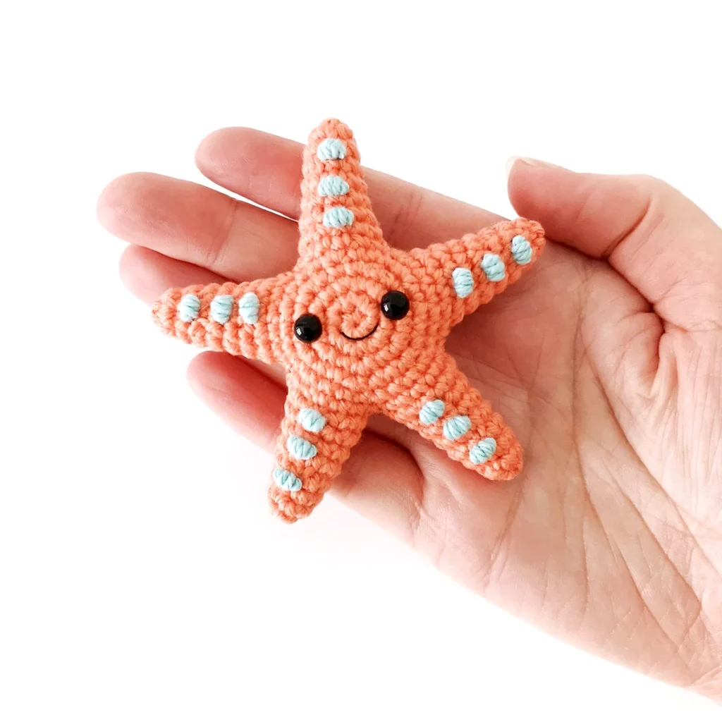 A small orange crocheted starfish with blue spots and a smiling face, lying on an open hand.