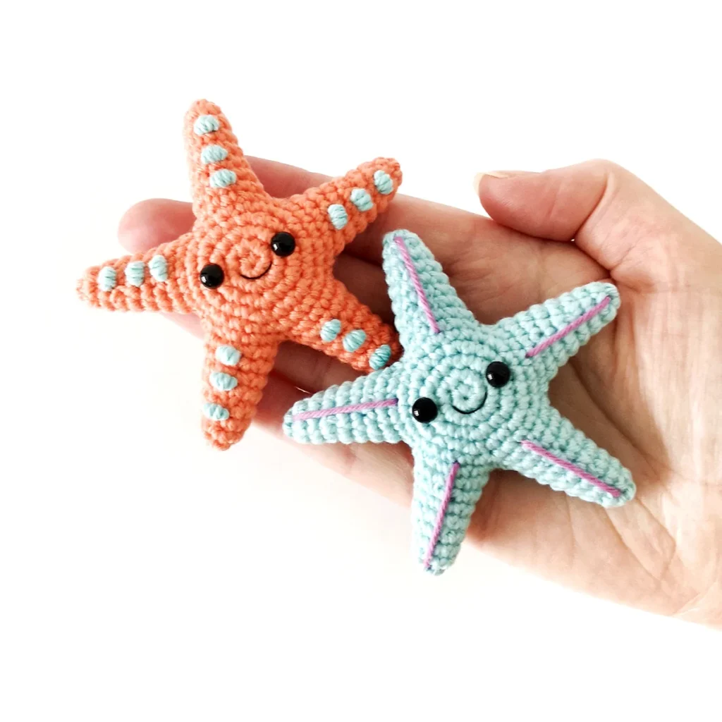 Two small crochet starfish, one orange with blue spots and one blue with purple lines, lying on an open hand.