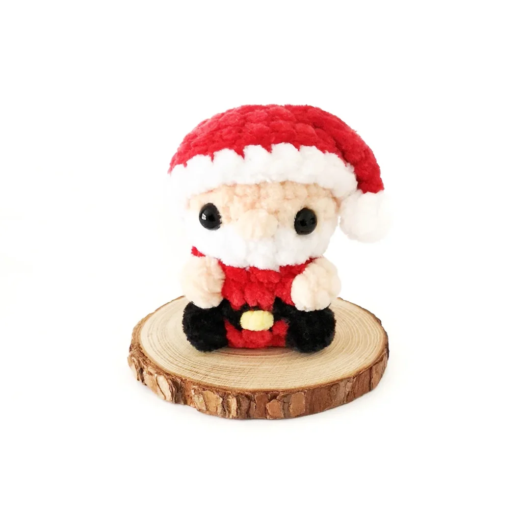 A small plush crochet Santa sitting on a round wood slice, against a white background.