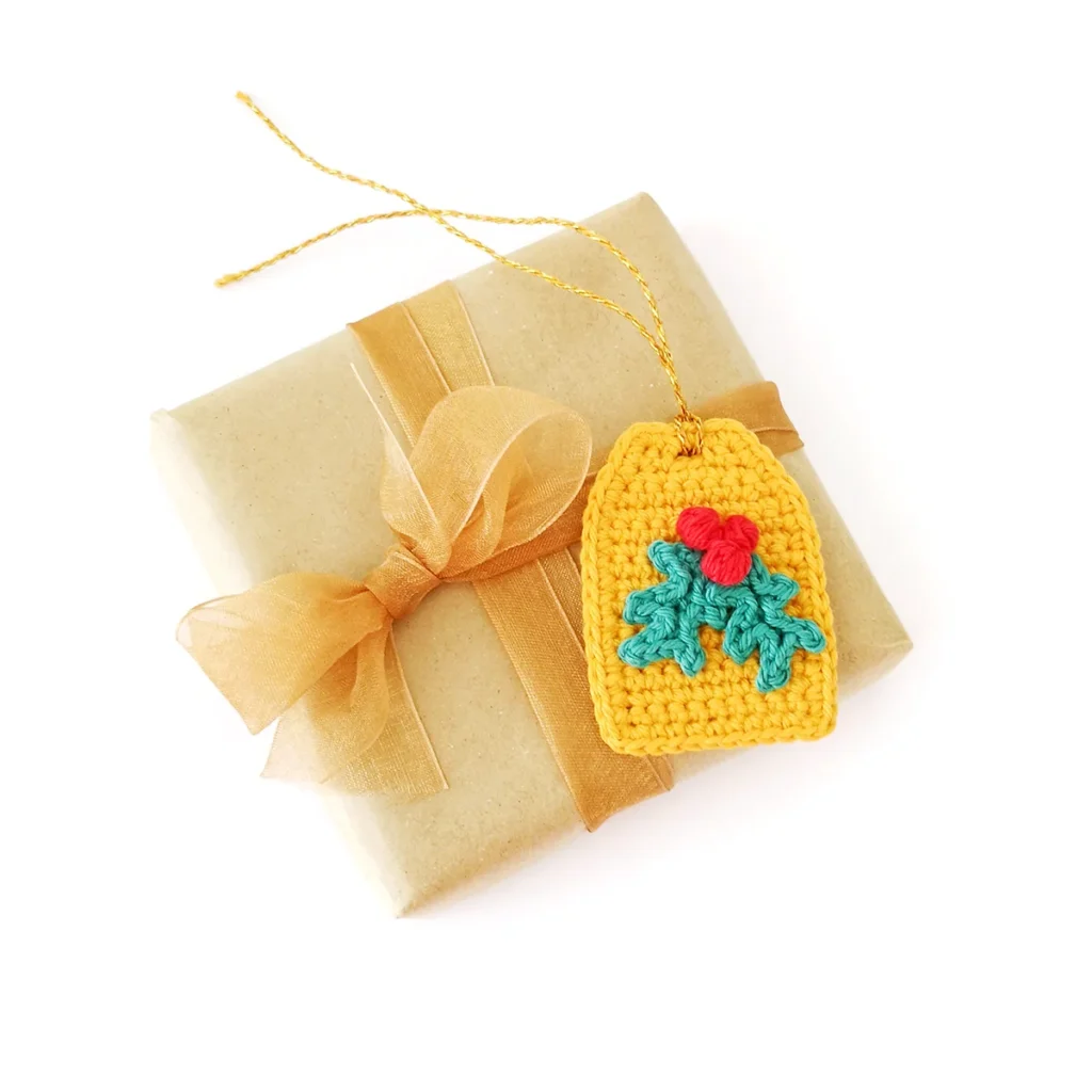 Crochet holly applique with two green leaves and a red berry cluster, attached to a yellow crochet gift tag with gold tie, lying on top of a brow paper wrapped gift.