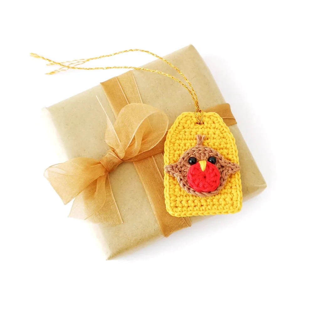 Crochet brown robin applique with red breast and yellow beak, attached to a yellow crochet gift tag with gold tie, lying on a brown paper wrapped gift.