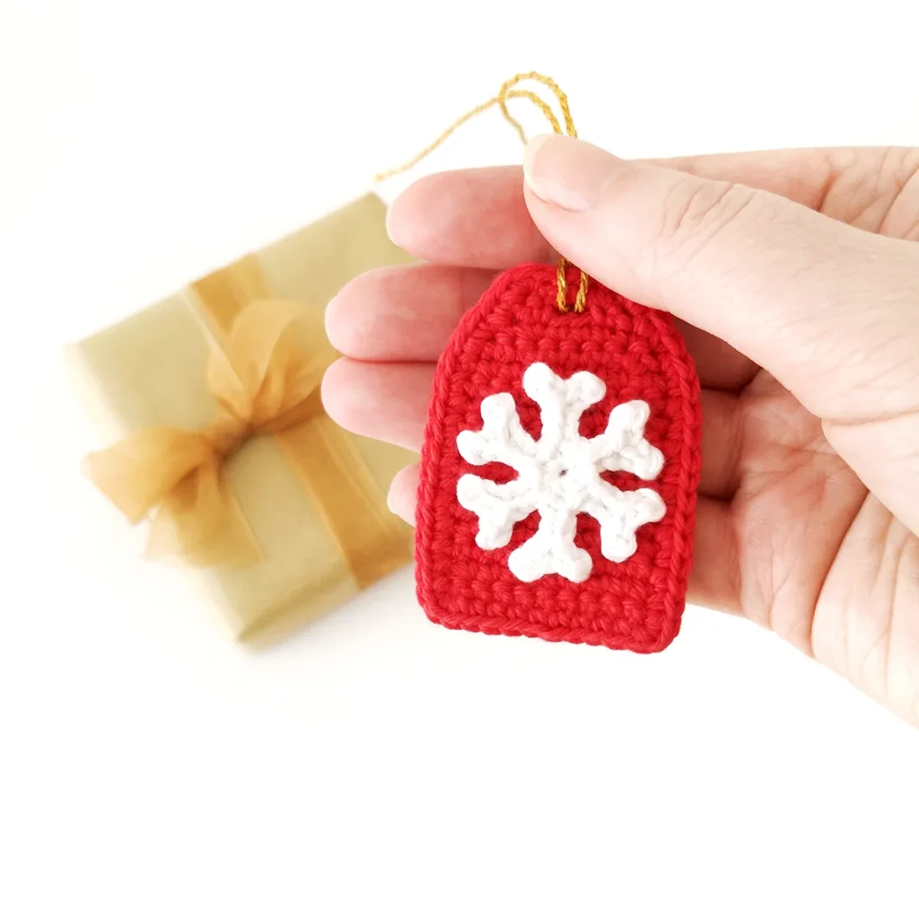 A crochet snowflake applique attached to a red crochet gift tag being held up by  a hand with a brown paper wrapped gift in the background.