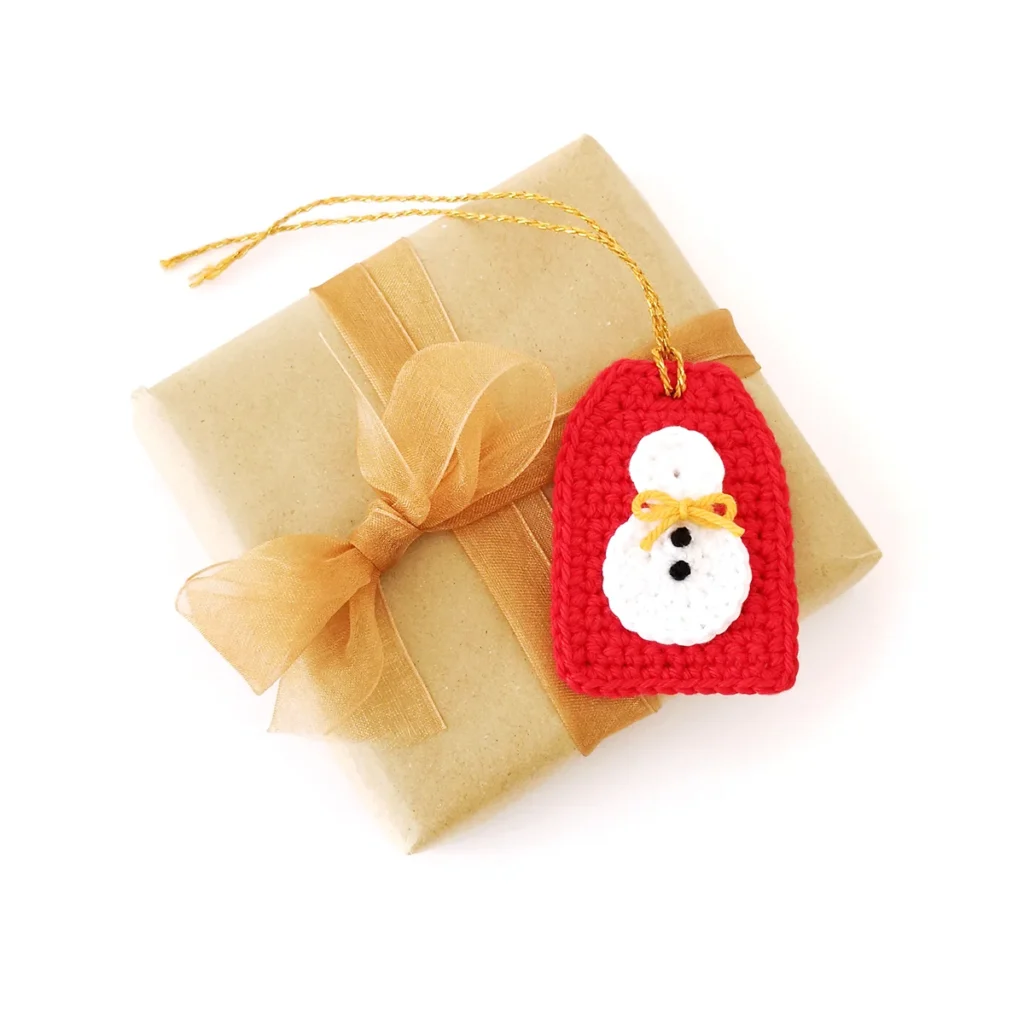 A crochet snowman applique with yellow bow and small black French knots for buttons, attached to a red crochet gift tag with gold tie, lying on top of a brown paper wrapped gift.