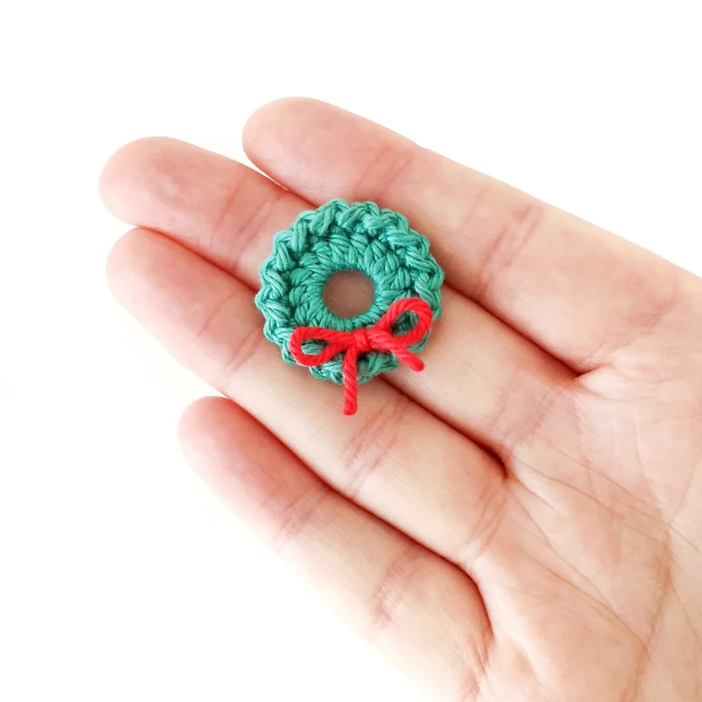 A green crochet Christmas wreath lying on the fingers of a hand against  a white background.