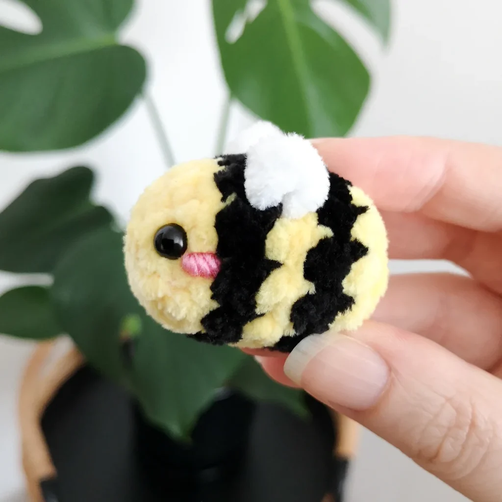 A small plush crochet bee with pink cheek being held up in front of a green plant.