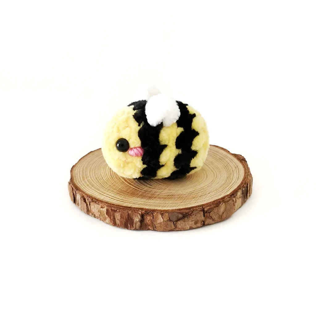 A small plush crochet bee with pink cheek sitting on a round slice of wood against a white background.