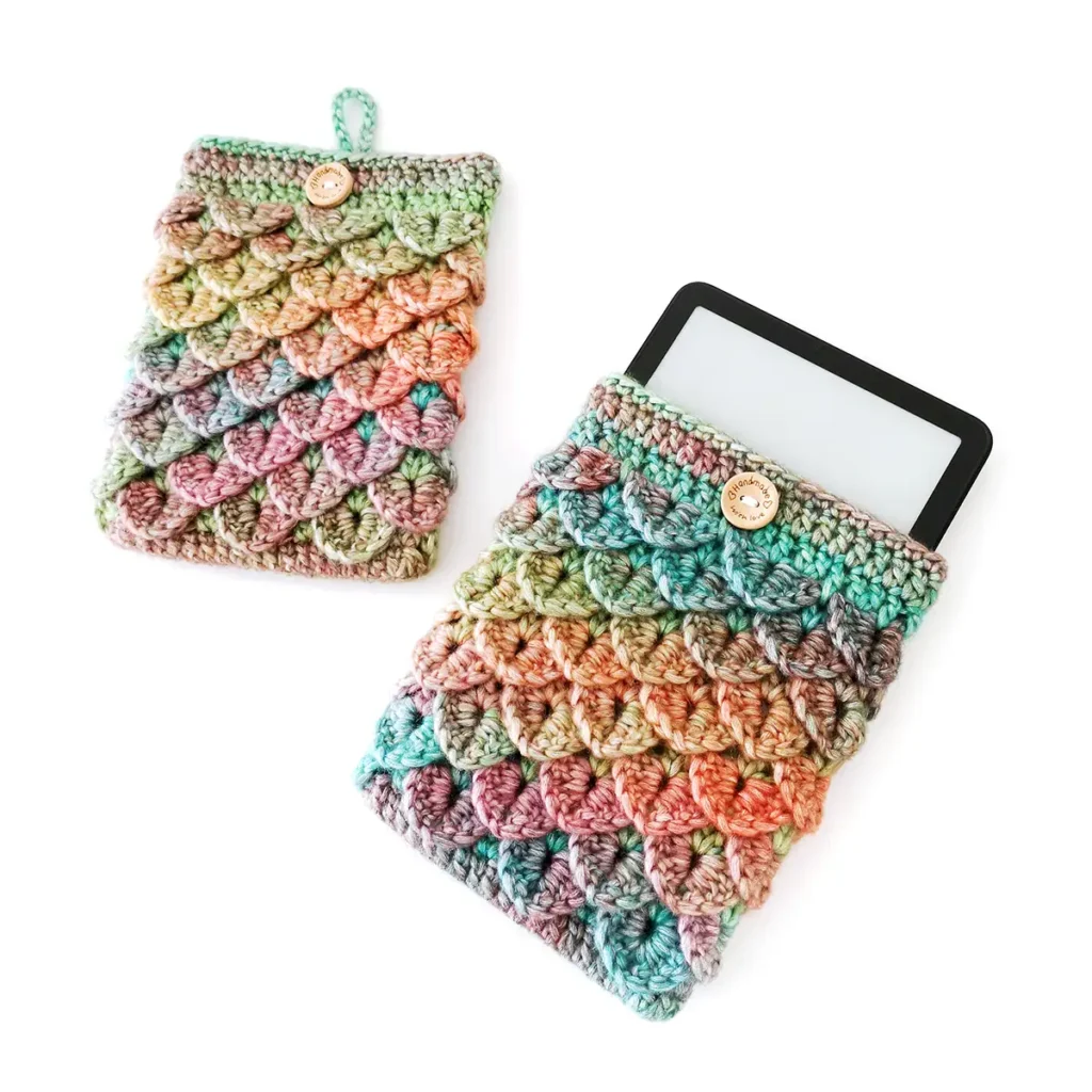 Two blue, green, orange and purple gradient crochet kindle sleeves lying side by side on a white background, featuring a textured dragon scale stitch pattern and a button loop closure, with a kindle peeking out the top of one of them.