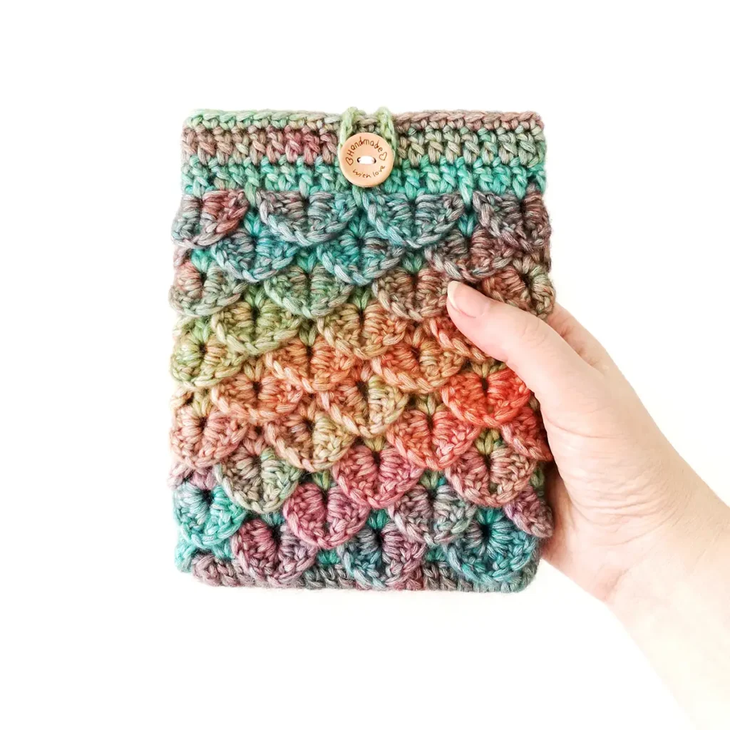 A blue, green, orange and purple gradient crochet kindle sleeve being held up against a white background, featuring a textured dragon scale stitch pattern.