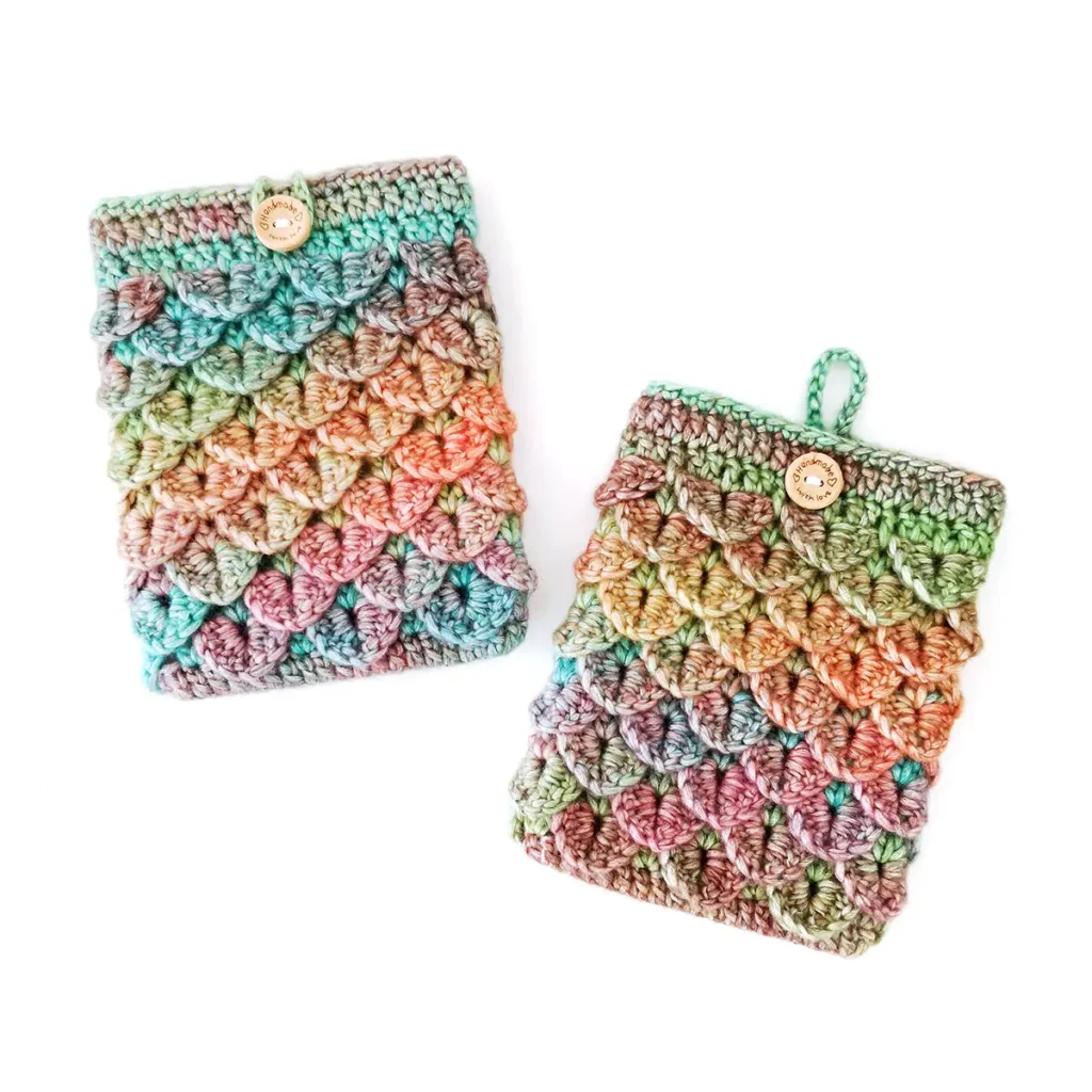 Two blue, green, orange and purple gradient crochet kindle sleeves lying side by side on a white background featuring a textured dragon scale stitch pattern and button loop closure.