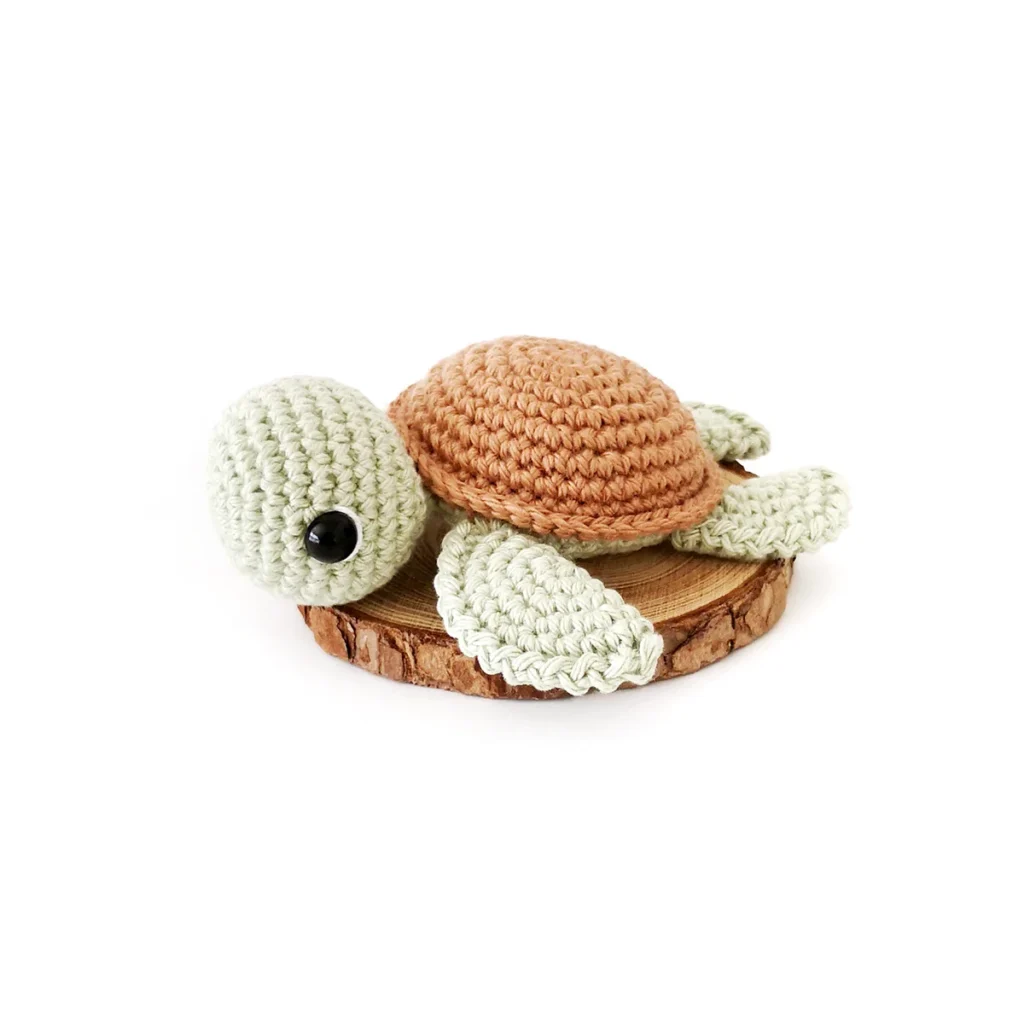 A small pale green crochet turtle amigurumi with brown shell lying flat on a wood round against a white background.