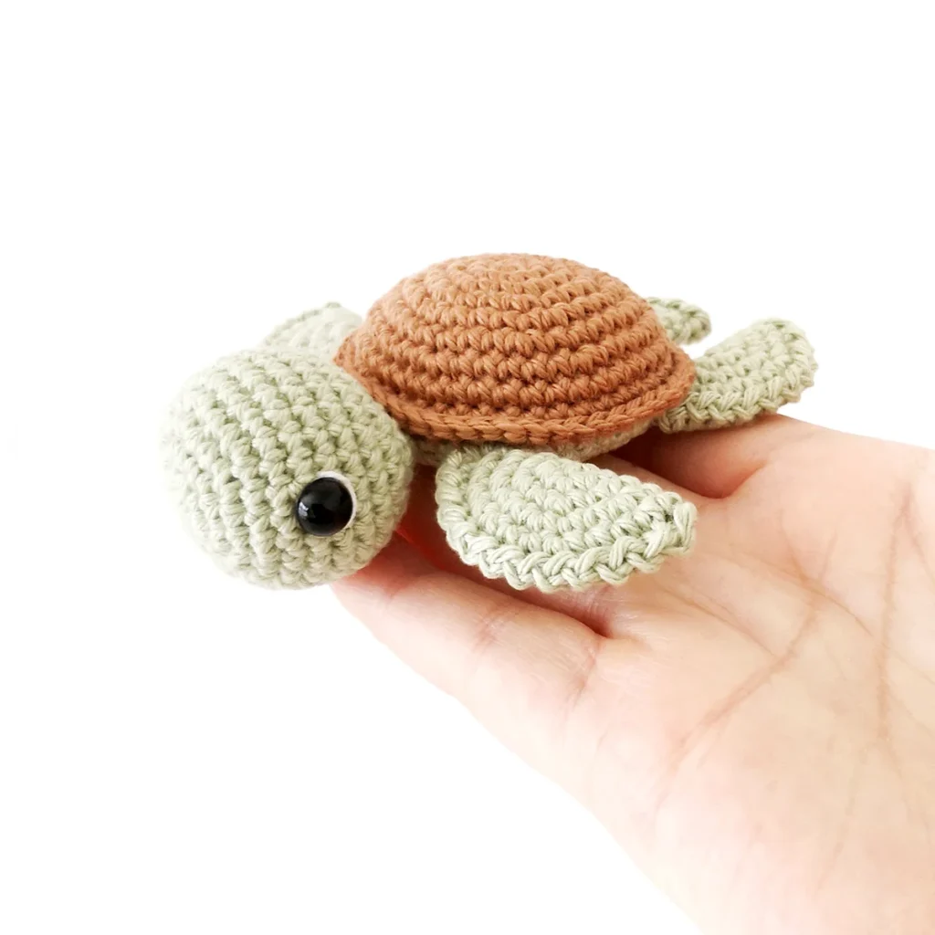 A small pale green crochet turtle amigurumi with brown shell lying flat on the end of a hand against a white background.