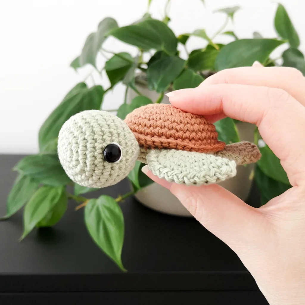 A small pale green crochet turtle amigurumi with brown shell being held up in front of a green trailing plant.