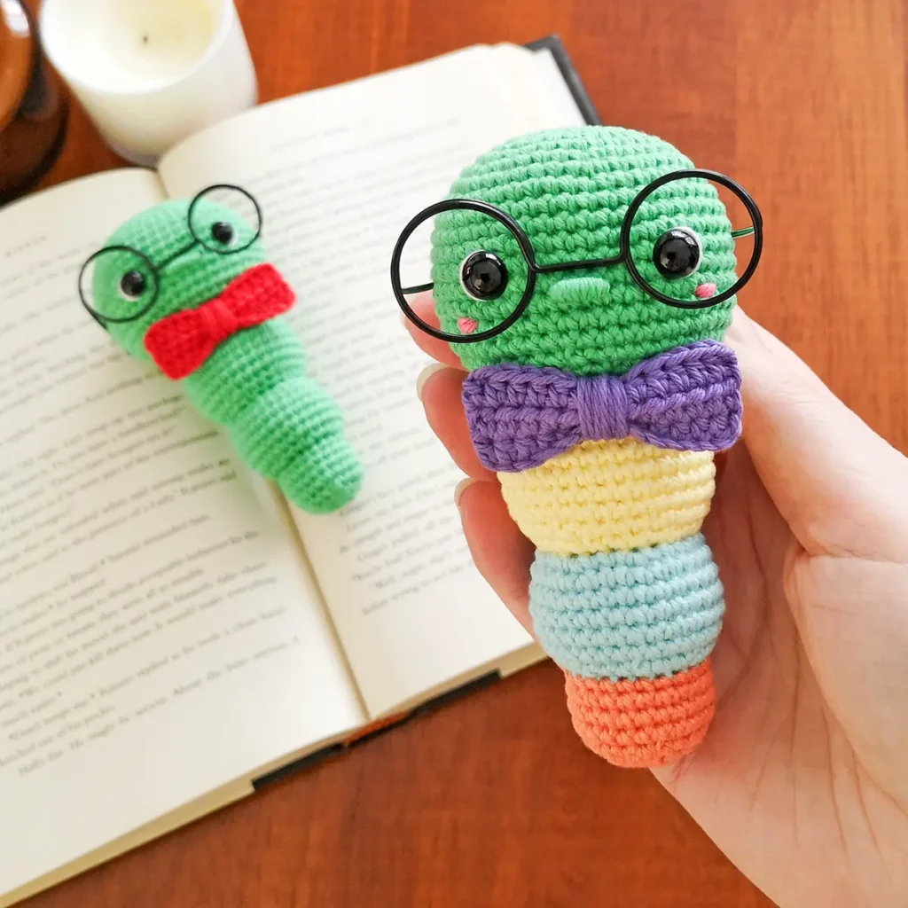 A crochet book worm with green head, yellow, blue and orange body segments, a purple bow tie and oversized black rimmed glasses being held in a hand with a green crochet bookworm lying on an open book in the background.