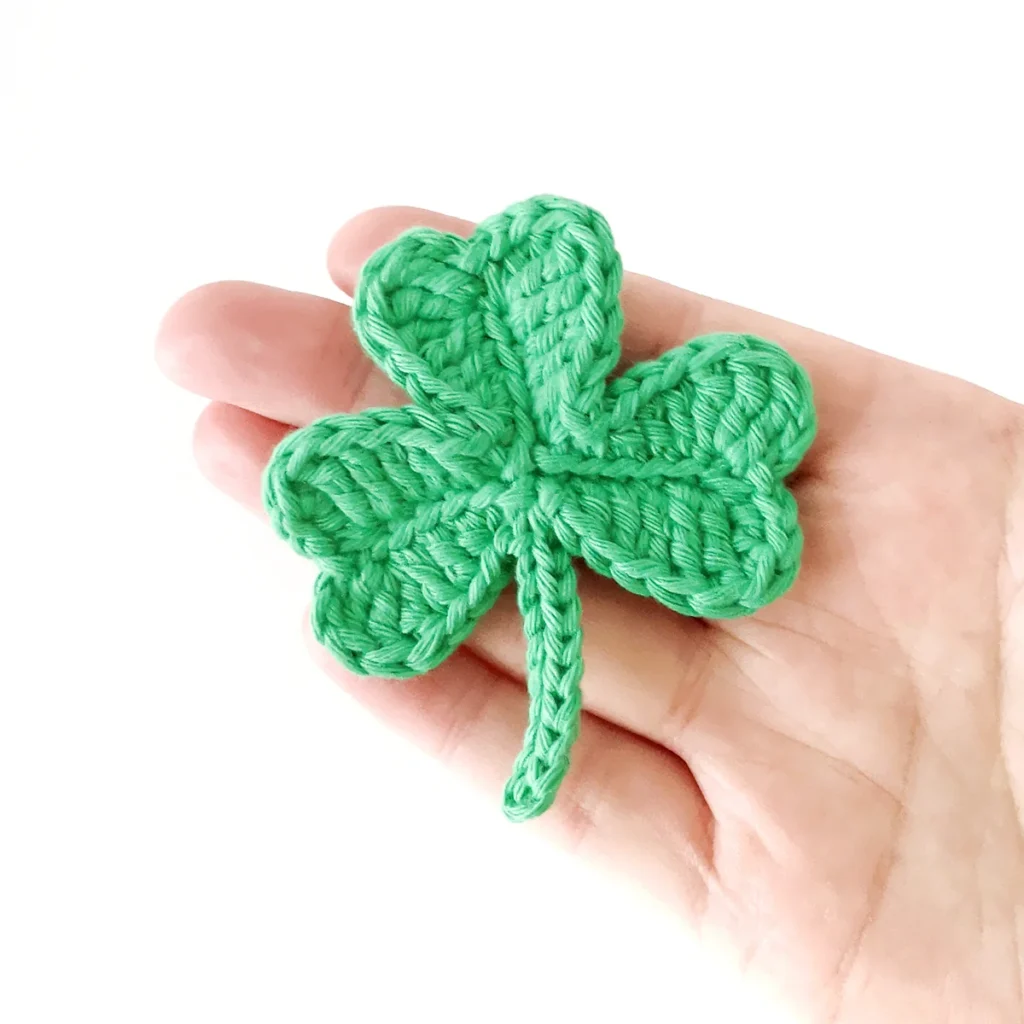 A green crocheted shamrock resting on the end of a hand against a white background.