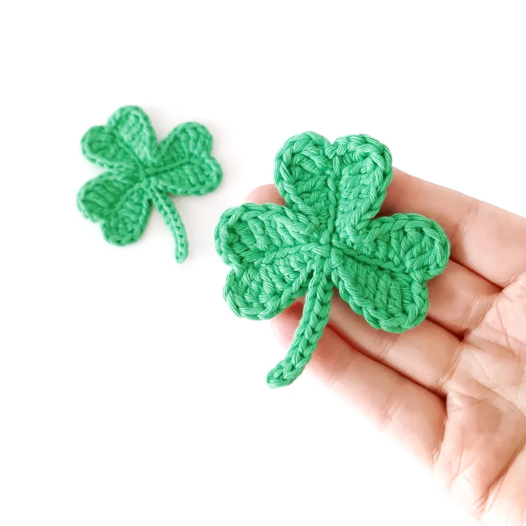A green crocheted shamrock resting on the end of a hand against a white background with another crochet shamrock in the background.
