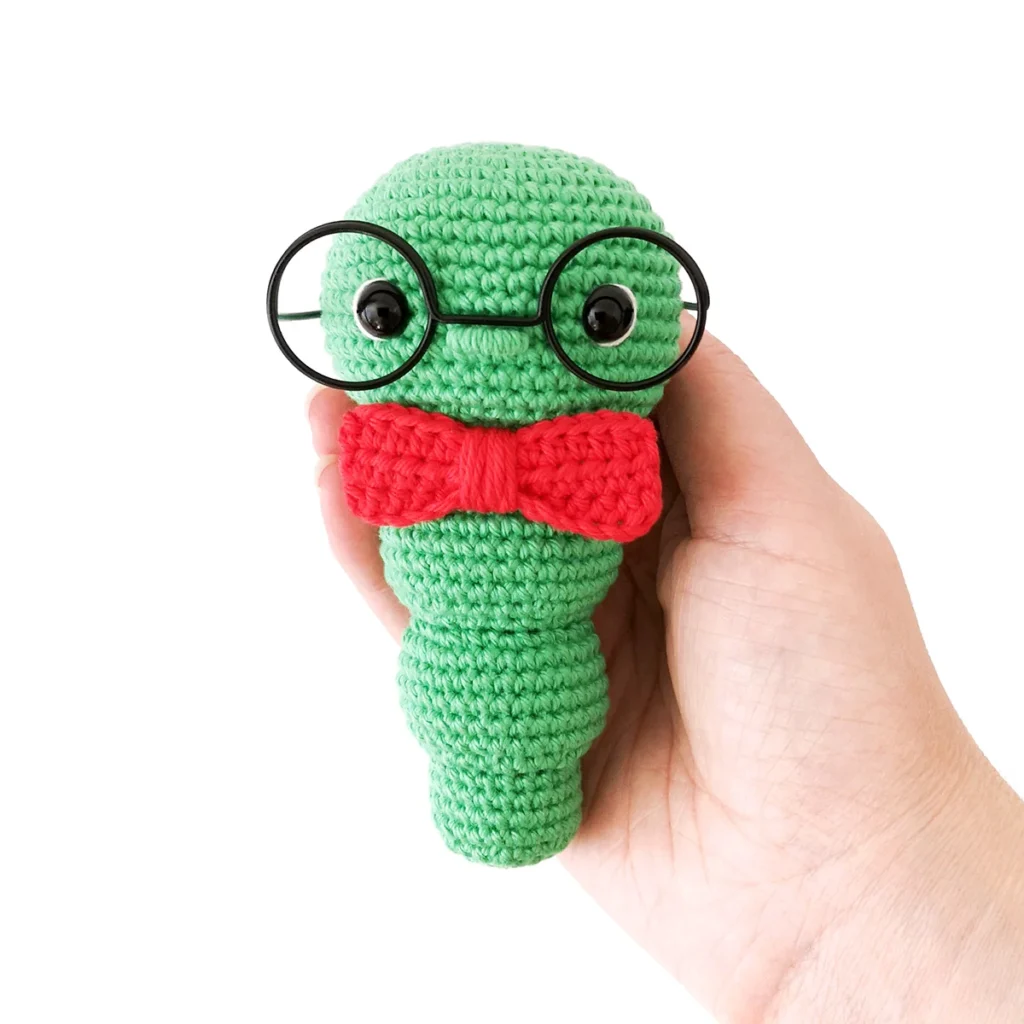 A green crochet bookworm with red bow tie and oversized black rimmed glasses being held in a hand against a white background.