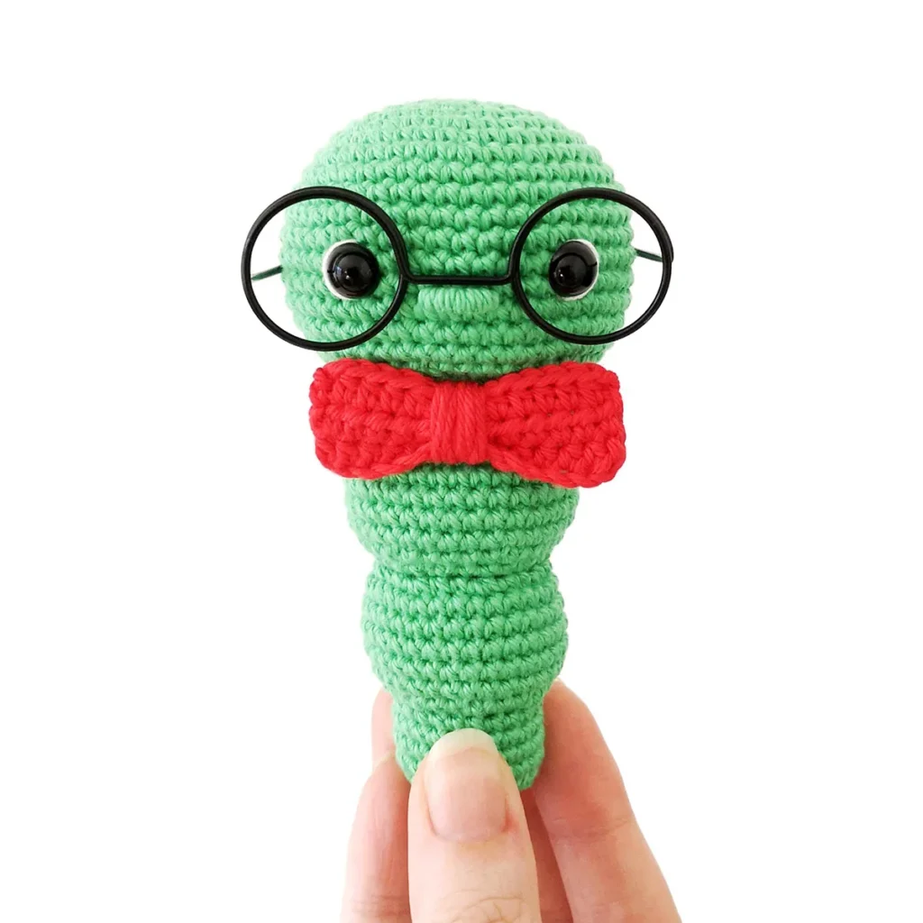 A green crochet bookworm with red bow tie and oversized black rimmed glasses being held at the bottom by a hand against a white background.