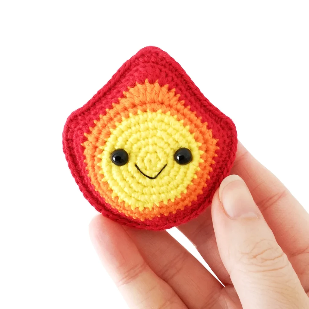 A small crochet fire with a smiling face and colours changing from yellow, orange and red, being held up by a hand.
