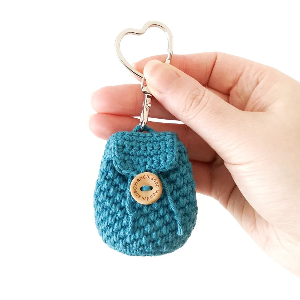 A blue mini crochet coin pouch keychain with textured body and button closure, being held up by a hand.