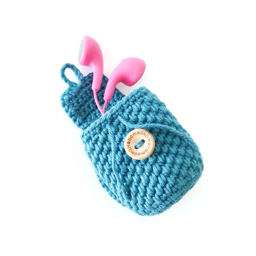 A blue mini crochet coin pouch keychain with textured body and button closure, lying open with a pair of pink earbuds sticking out.