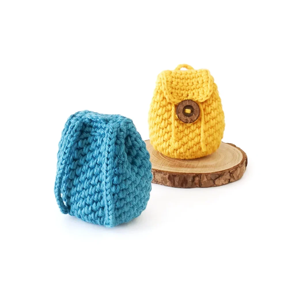 Two mini crochet backpack coin pouches, one blue and one yellow. The yellow coin pouch is sitting on a small wood round and facing forwards with the blue coin pouch sitting in front with the back facing forward.