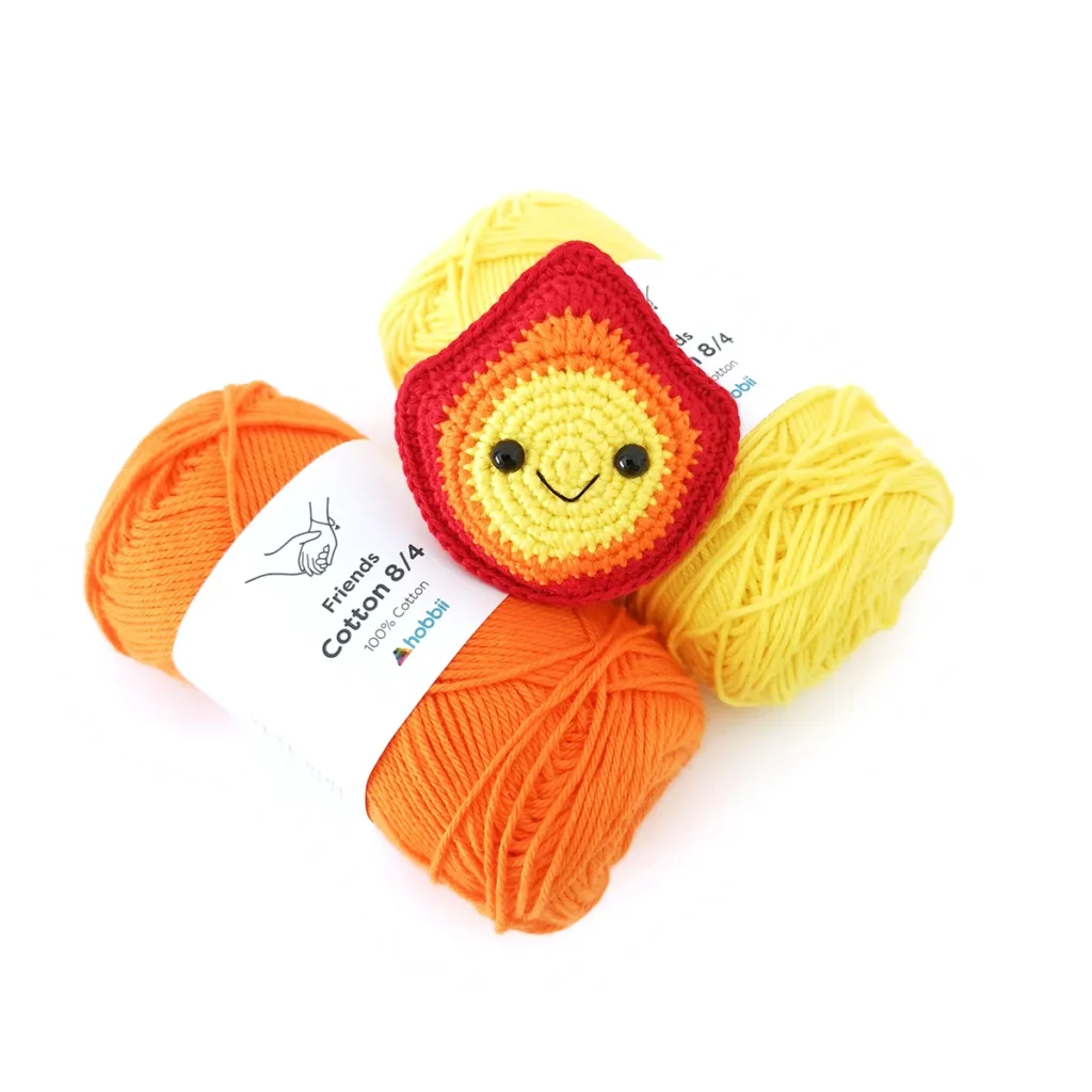 A small crochet fire with a smiling face and colours changing from yellow, orange and red, lying on a yellow and orange skein of yarn.