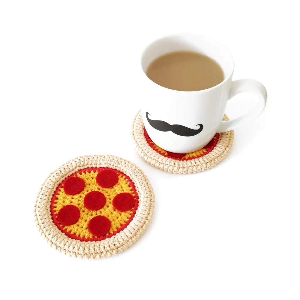 Two crochet pizza coasters lying on a white background, one has a mug with a moustache motif sitting on it.
