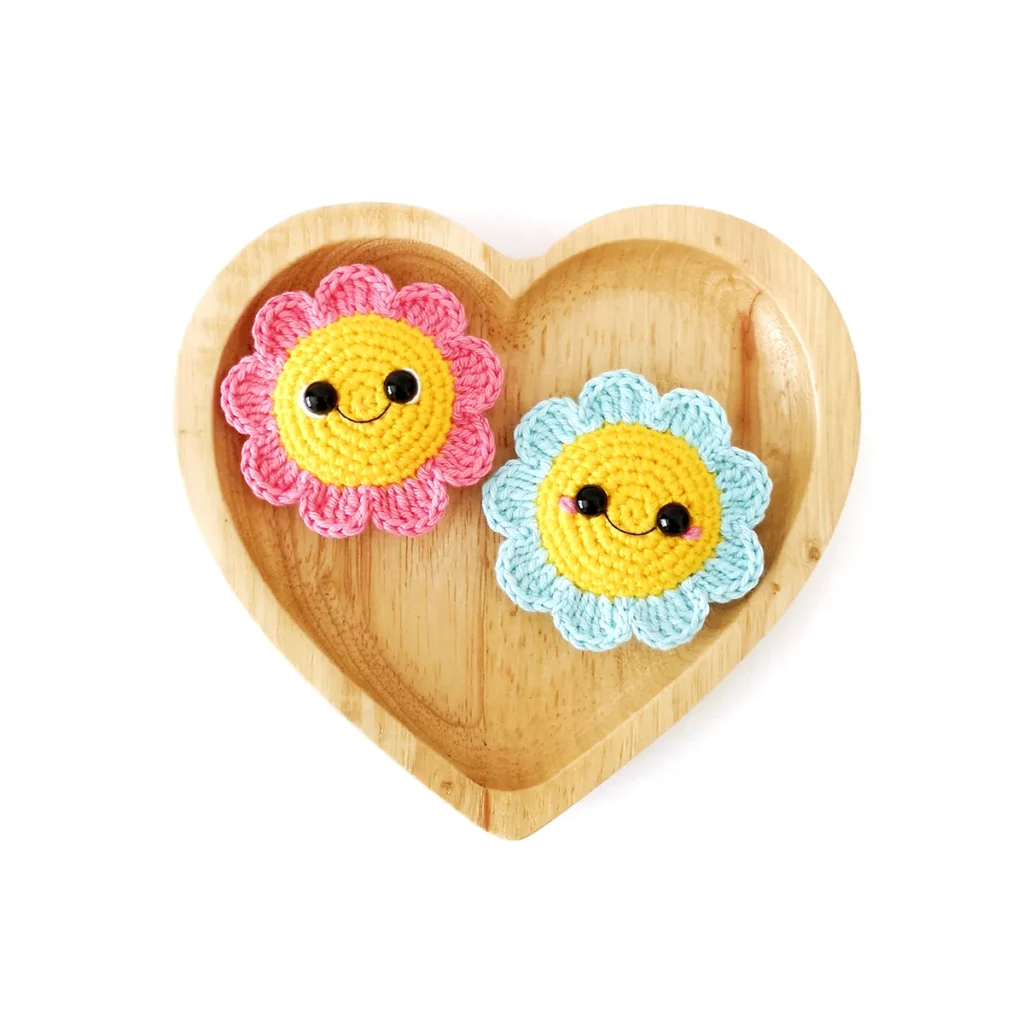 Two amigurumi flowers, one light blue and one pink, with yellow centre and smiling face, lying flat on a wooden heart shaped tray.