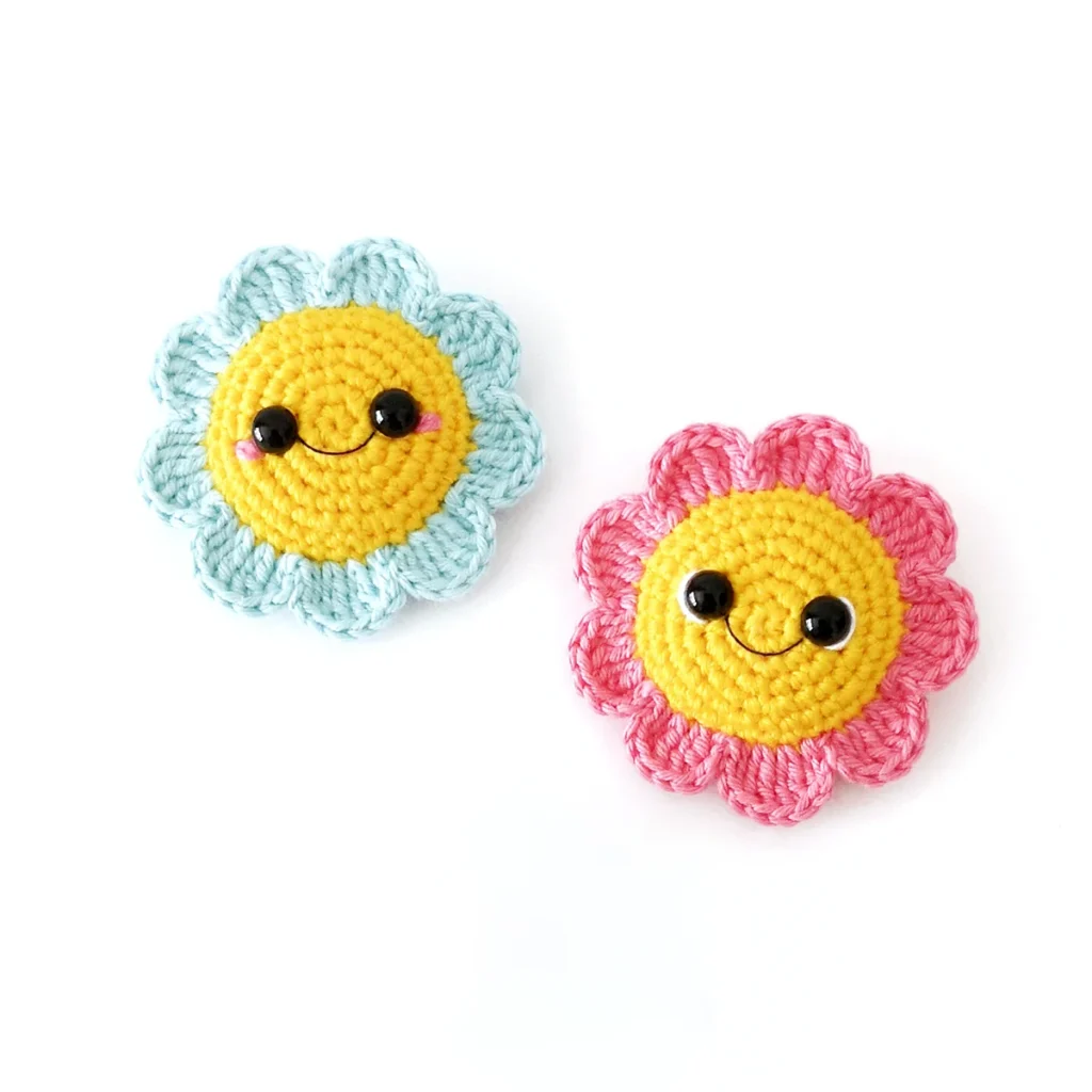 Two amigurumi flowers, one light blue and one pink, with yellow centre and smiling face, lying on a white background.