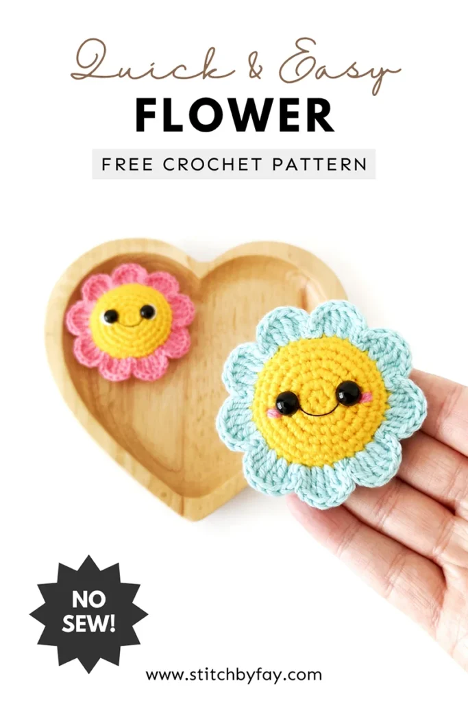 Pinterest image for a crochet flower pattern. Image shows a light blue crochet amigurumi flower with yellow centre and smiling face, lying flat on the end of a hand with a pink crochet flower lying on a heart shaped wooden tray in the background. Text overlay reads: "Quick & easy flower, free crochet pattern, www.stitchbyfay.com" with "no sew!" in a starburst.